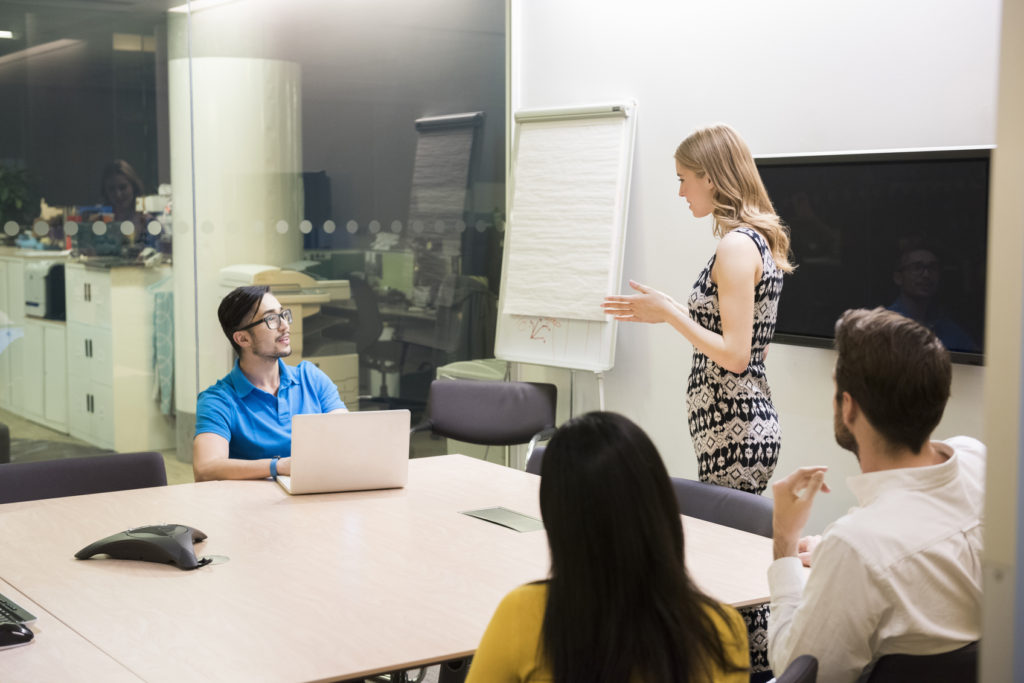 A photo of businesswoman giving presentation to colleagues at conference table. Business people are in board room. Concentrated associates listening to presentation, in a brightly lit modern office.