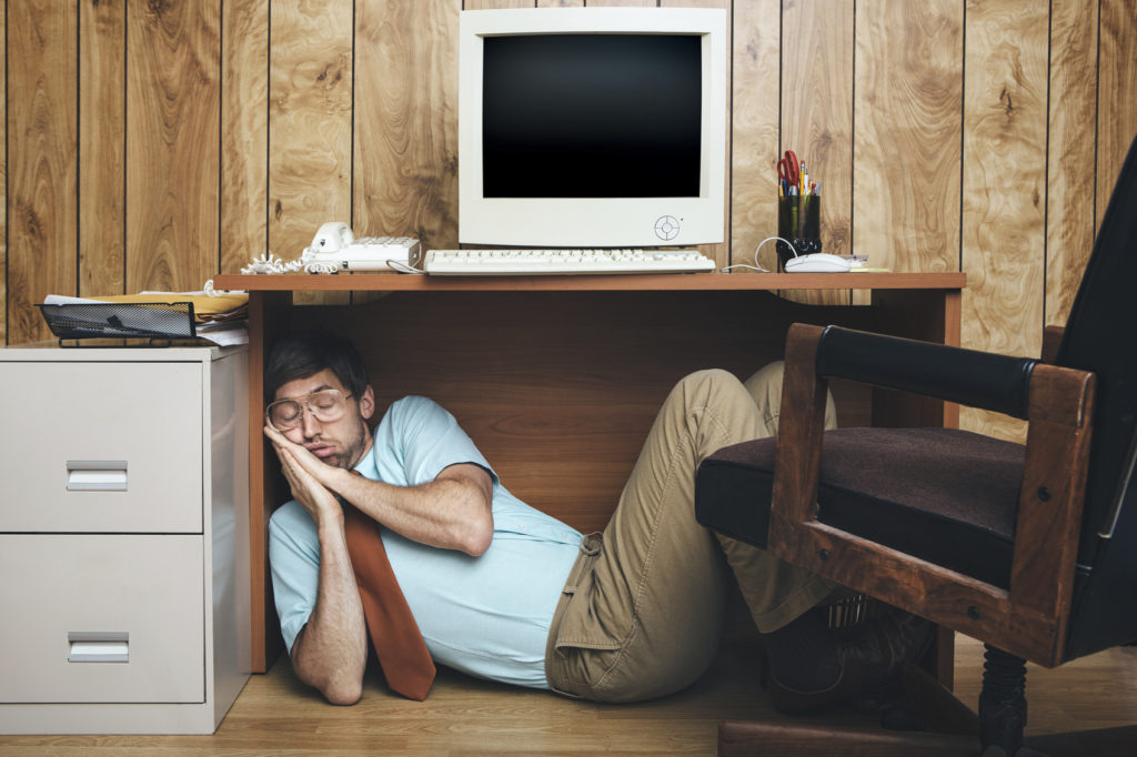 A man and office in 1980's - 1990's style, complete with vintage computer and technology of the time, sleeps under his desk, too tired and bored to continue working. Wood paneling on the wall in the background.